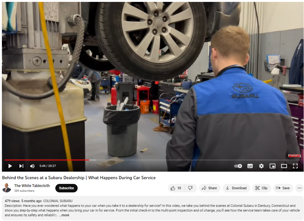Screenshot from a YouTube video showing a mechanic at a Subaru dealership, viewed from behind, overseeing a car lifted on a hoist for maintenance. The busy garage setting includes tools and another mechanic in the background.